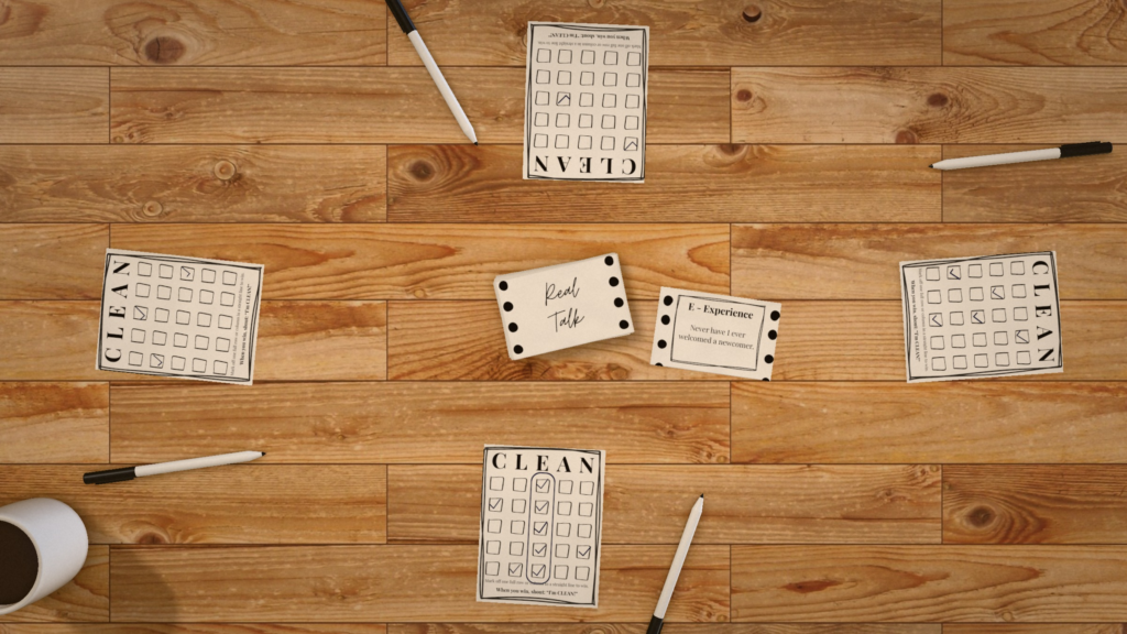 Overhead view of a CLEAN recovery game for four players: a face-down Real Talk pile and drawn card in the center, with a CLEAN board and pen at each seat and a coffee mug